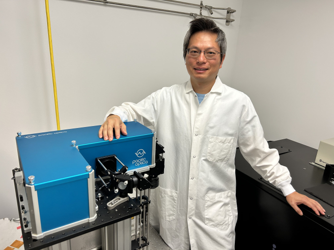 Che-Hang Yu stands in white lab coat in front of Pacific Optica technology